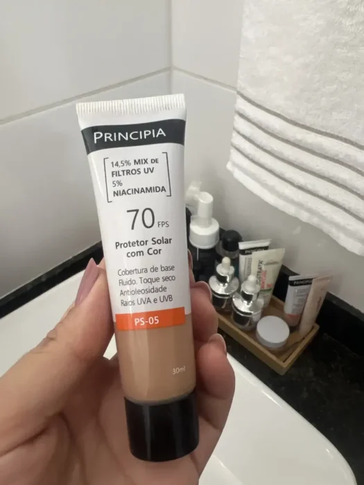 Minha mão segurando o Protetor Solar Principia com Cor PS-05 no banheiro.