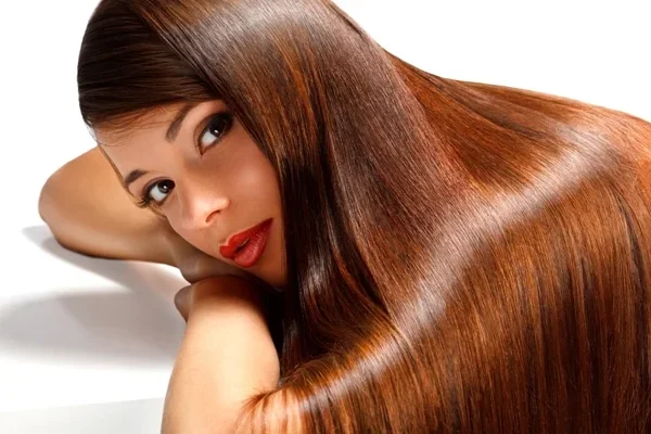 Mulher com cabelo longo castanho acobreado super liso e com brilho espelhado, deitada de braços cruzados, exemplificando as tendências de haircare para 2026.