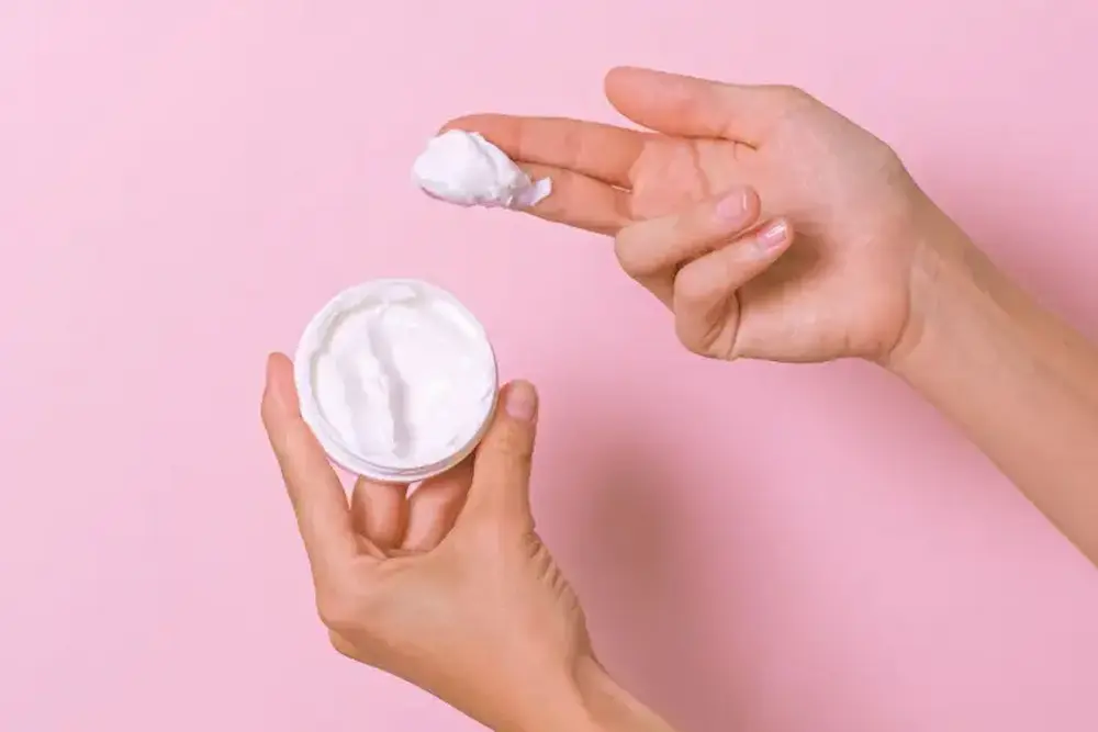 Mão pegando creme hidratante de um pote com fundo rosa para explicar a função da barreira cutânea.
